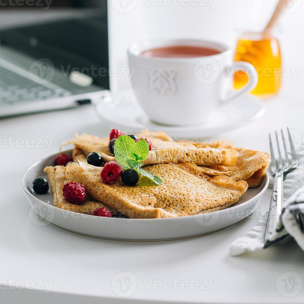 delicioso café da manhã. mesa de mesa branca com laptop e panquecas de crepes frescas, xícara de chá e pote de mel. conceito de freelancer. comida de café da manhã saudável. foto