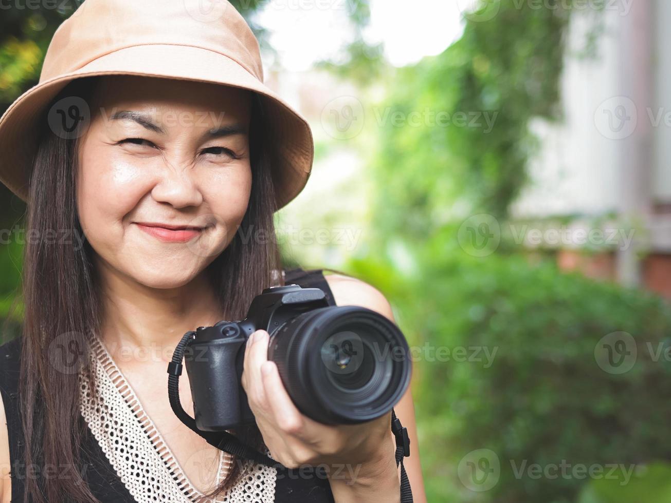 mulher asiática, usando chapéu e top preto sem mangas, de pé no jardim e segurando a câmera dslr, sorrindo alegremente. foto