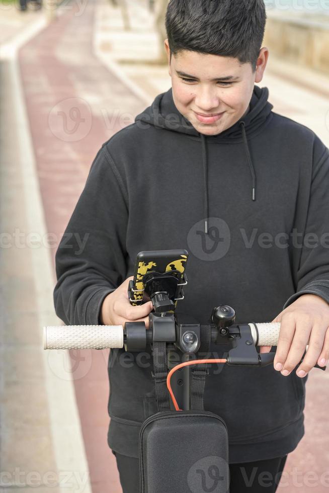 vista frontal 14-17 anos sorrindo alegremente andando de scooter elétrica na ciclovia e usando aplicativos de música para smartphones, conceito de lazer urbano de mobilidade elétrica foto