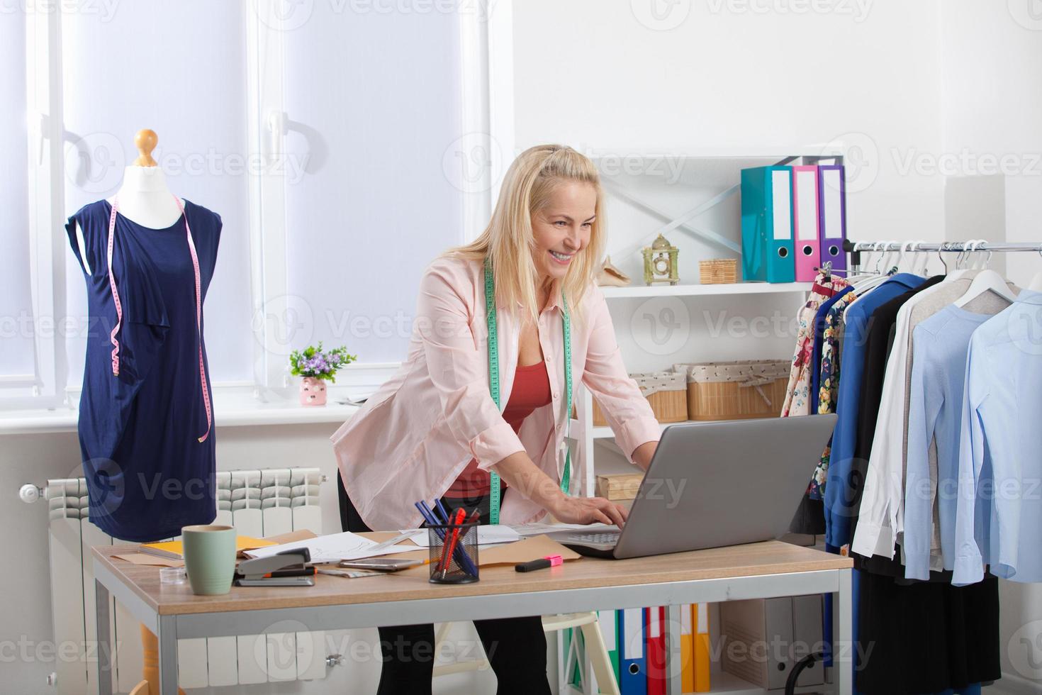 discussão do projeto de design de roupas com o cliente por telefone, skype. linda mulher europeia falando ao telefone em pé perto da mesa na oficina com roupas penduradas no fundo foto