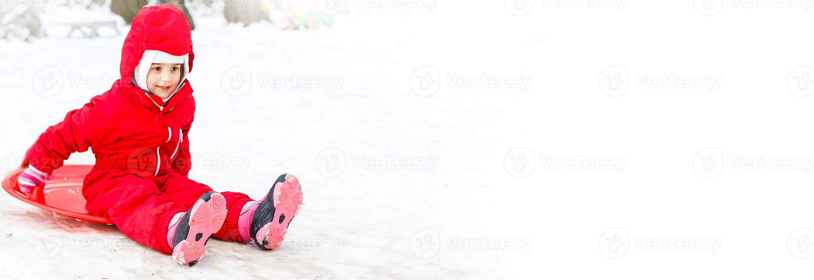 menina muito sorridente em seu traje de esqui deslizando por uma pequena colina coberta de neve com seu trenó foto