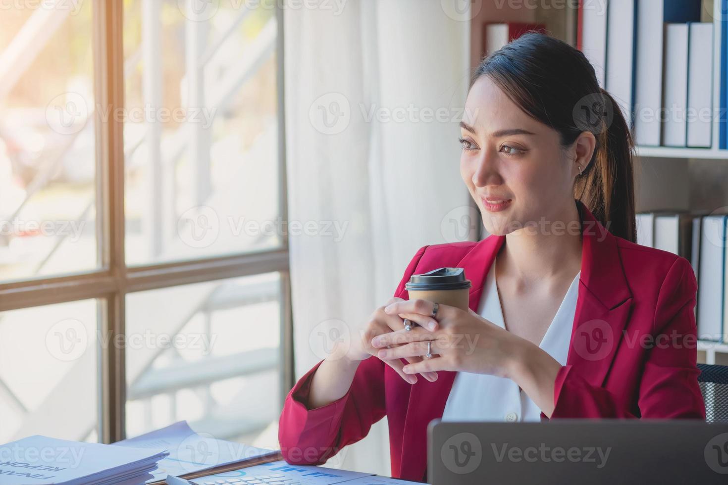 financeira, empresária asiática de terno vermelho segurando a xícara de café sentada na mesa no escritório, tendo computador para fazer trabalho de contabilidade no local de trabalho para calcular o lucro anual por dever, ideia de negócio foto