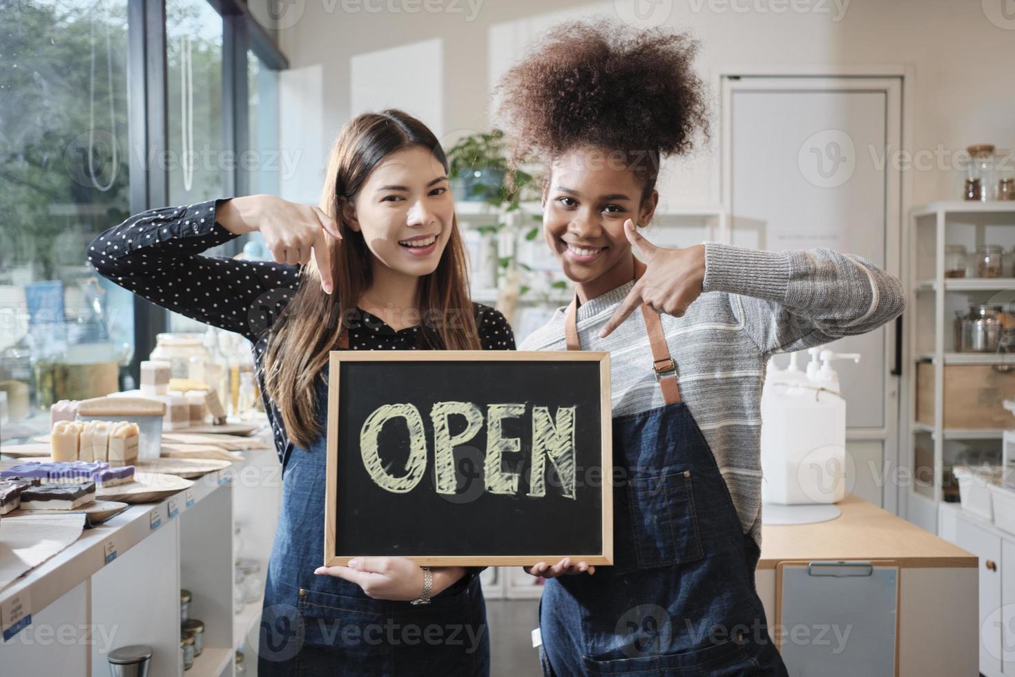 duas jovens lojistas mostram uma placa aberta com sorrisos alegres na loja de recarga, trabalho feliz com produtos orgânicos, mantimentos sem desperdício, mercadorias ecológicas e negócios sustentáveis. foto