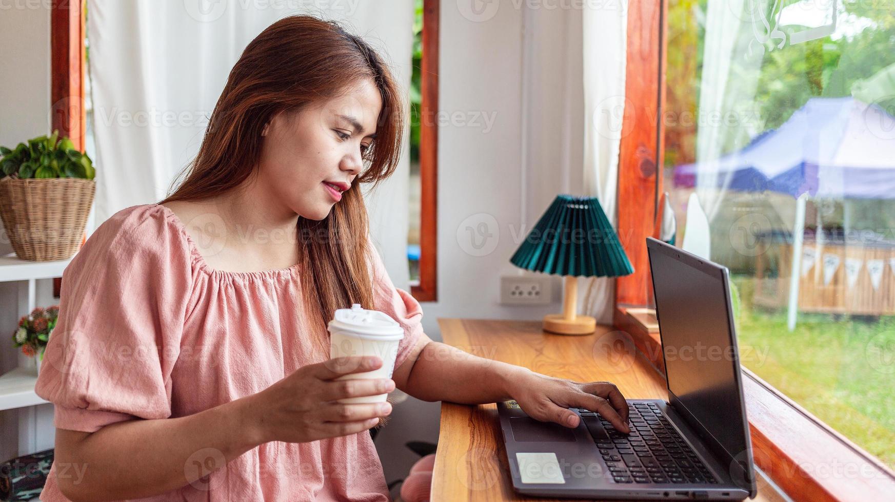 uma mulher feliz em um café usando um laptop na mão e uma xícara de café de papel. jovem mulher branca com cabelos longos, sentada em uma cafeteria ocupada trabalhando em seu laptop. foto