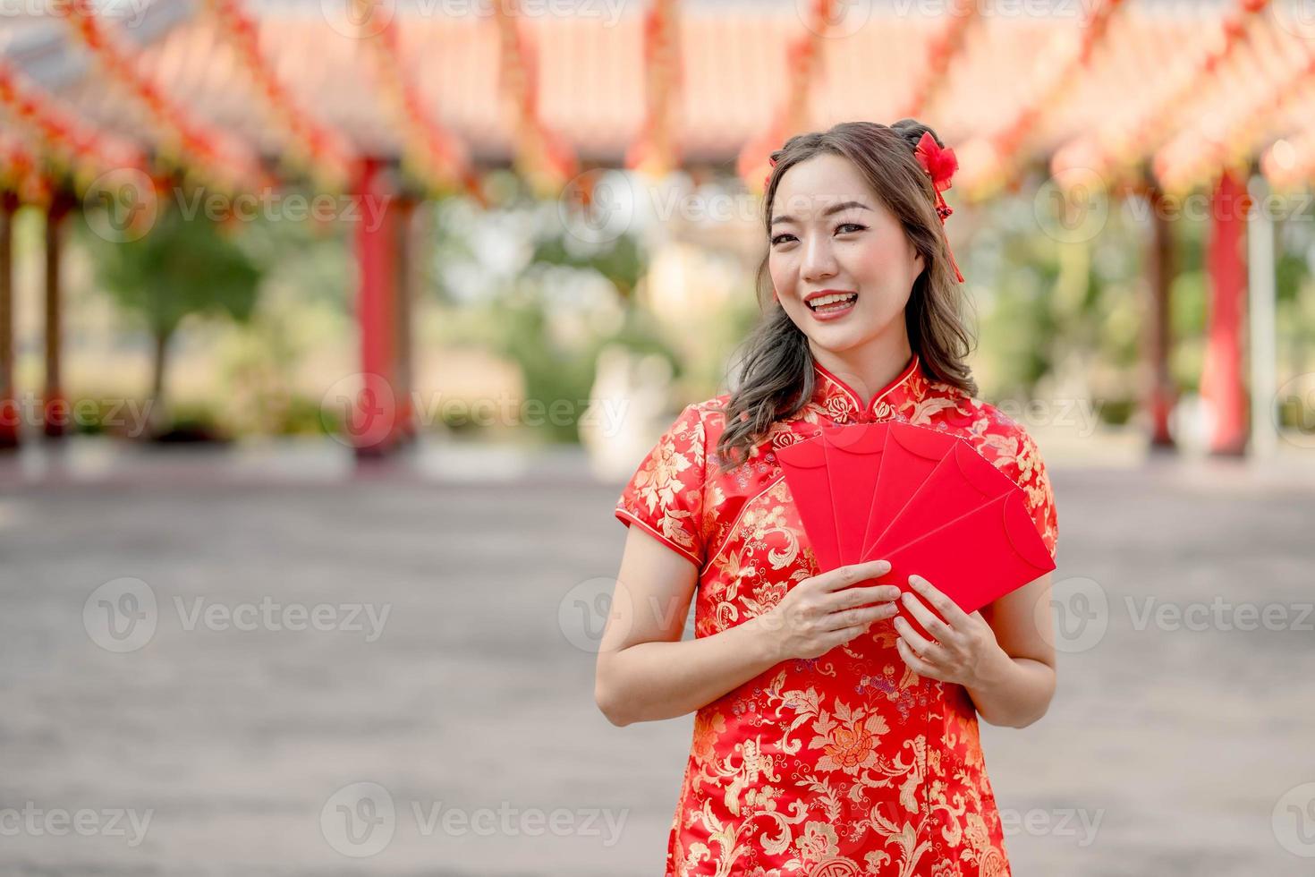 jovem mulher asiática sorrindo alegremente segurando ang pao, envelopes vermelhos vestindo cheongsam parecendo confiante no templo budista chinês. comemore o ano novo lunar chinês, feriado da época festiva foto