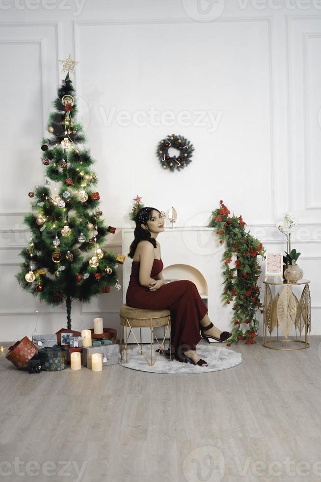 retrato de uma jovem bonita, confortável, sente-se, sorrindo, use um vestido vermelho na sala de estar decorada de natal dentro de casa foto