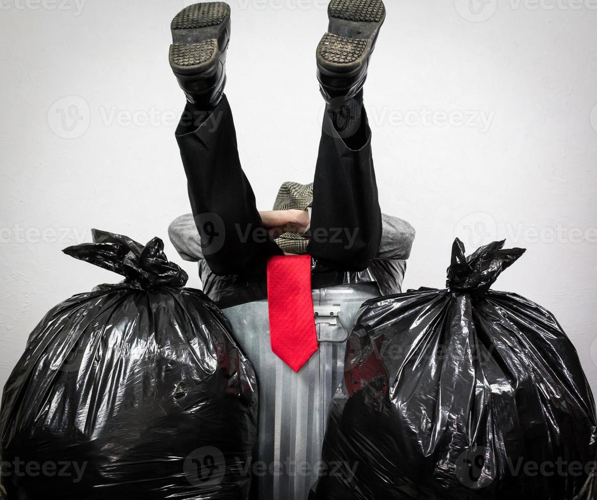 empresário de terno e chapéu e gravata vermelha sentado na lata de lixo cercado por sacos de lixo. deprimido. homem vencido pelo capitalismo e pela ganância. foto