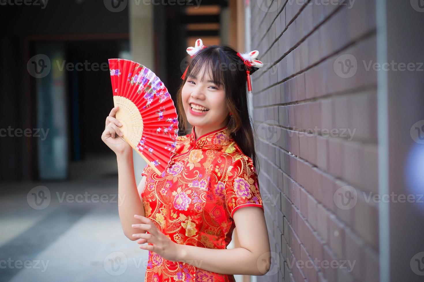 bela jovem asiática de vestido vermelho fica sorrindo alegremente olhando para a câmera segura um leque entre o antigo centro da cidade no tema do ano novo chinês. foto