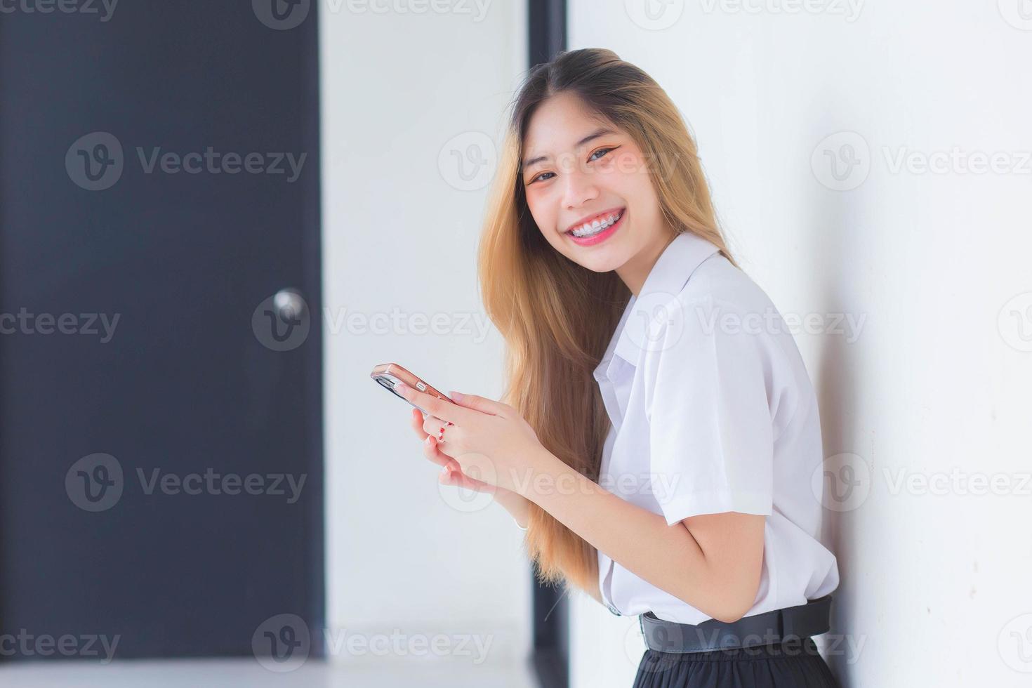 o retrato da estudante tailandesa asiática bonita de uniforme está sorrindo alegremente enquanto usa um smartphone na universidade. foto