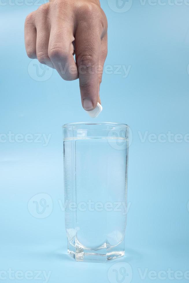 copo de vidro com água e uma mão com um comprimido na medicação de fundo azul foto