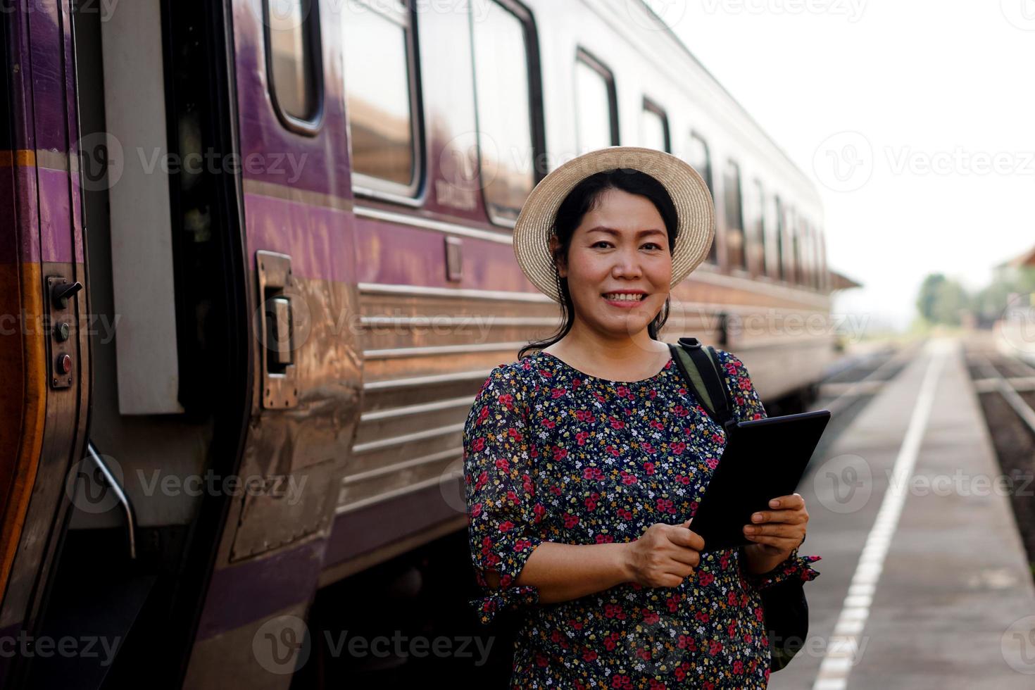 feliz viajante asiática de meia-idade está na estação ferroviária, usa chapéu, segura tablet inteligente e sorri. conceito, viajar de trem na tailândia pode reservar ou por bilhete online. foto