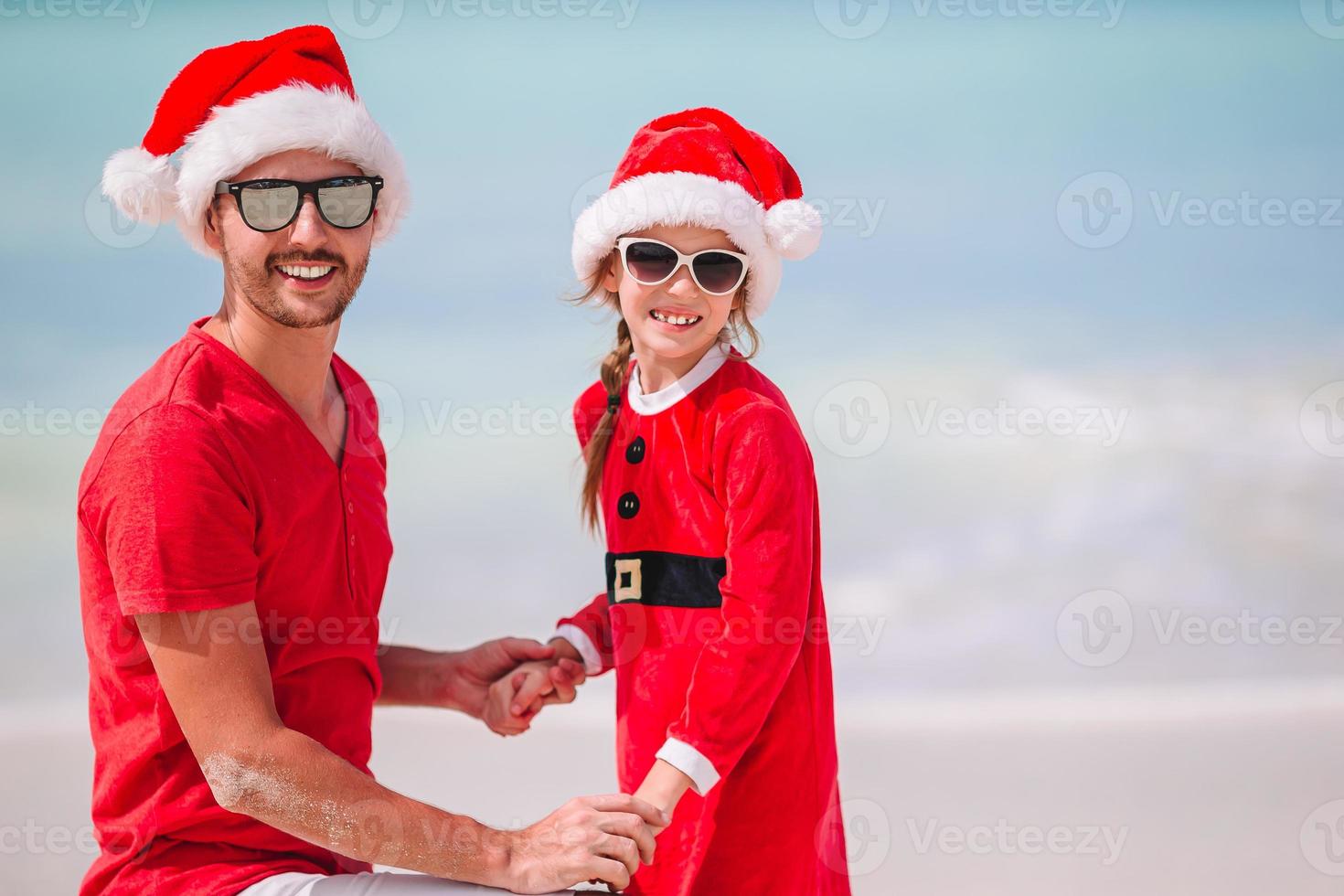 pai e filha com chapéu de Papai Noel se divertem na praia tropical foto