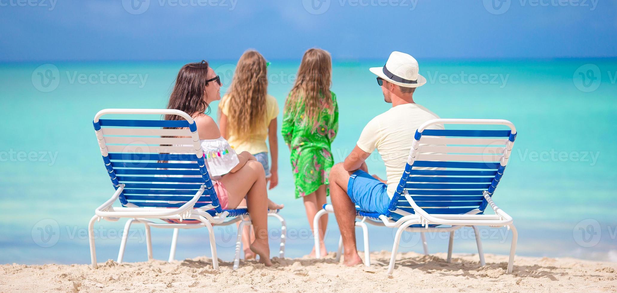 feliz linda família de quatro pessoas na praia. pais relaxando na espreguiçadeira e crianças se divertindo na costa foto