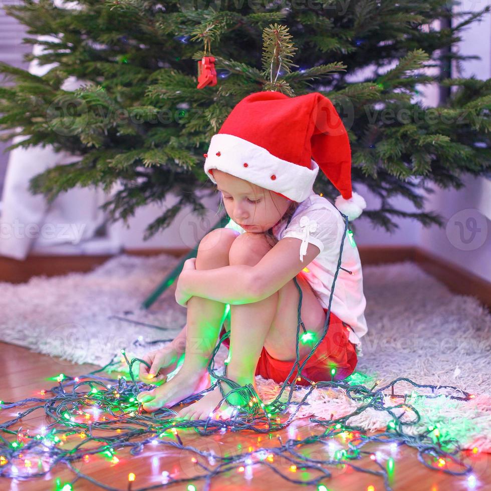 menina bonita com chapéu de papai noel sentado debaixo da árvore de natal entre guirlandas foto