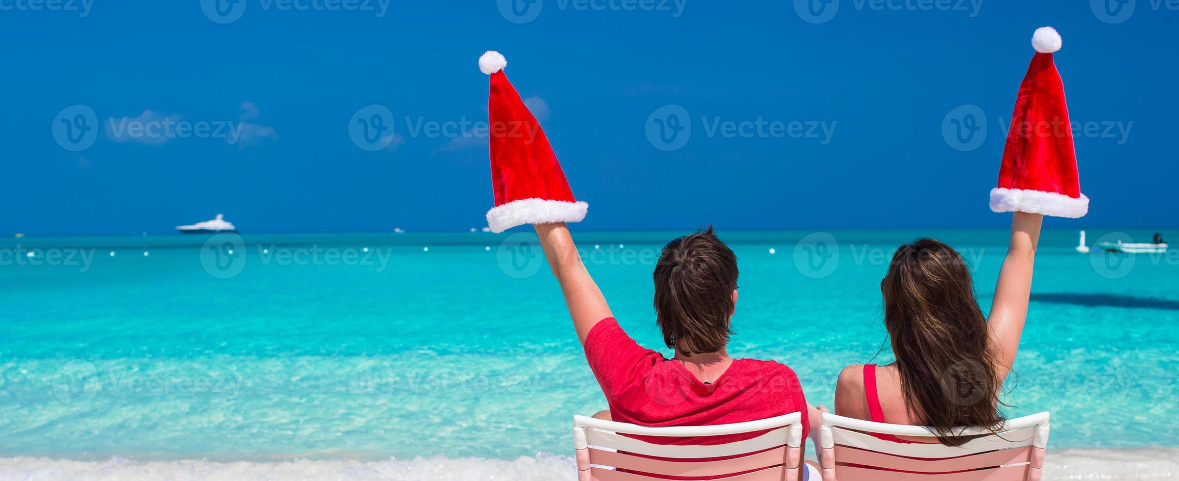 casal jovem feliz em chapéus de papai noel vermelhos sentados em cadeiras de praia foto