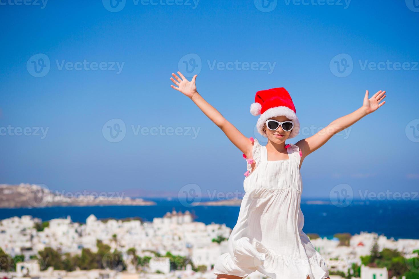 menina engraçada no chapéu vermelho de papai noel ao ar livre fundo de mykonos. garoto na rua da típica vila tradicional grega com paredes brancas e portas coloridas nas férias de natal na grécia foto