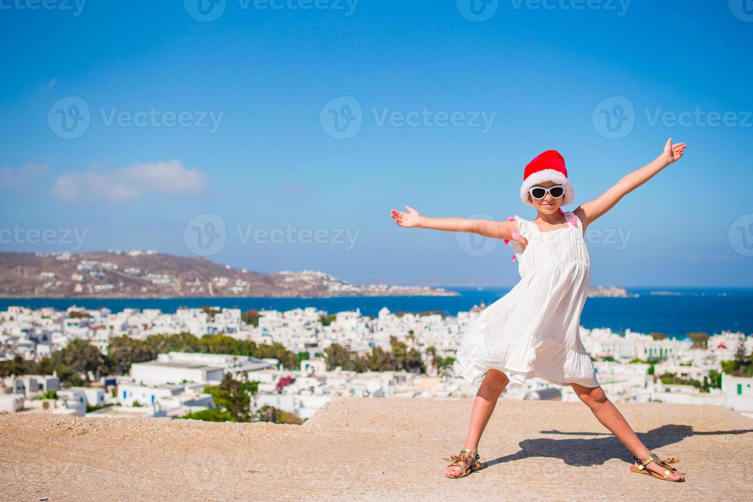 menina engraçada no chapéu vermelho de papai noel ao ar livre fundo de mykonos. garoto na rua da típica vila tradicional grega com paredes brancas e portas coloridas nas férias de natal na grécia foto