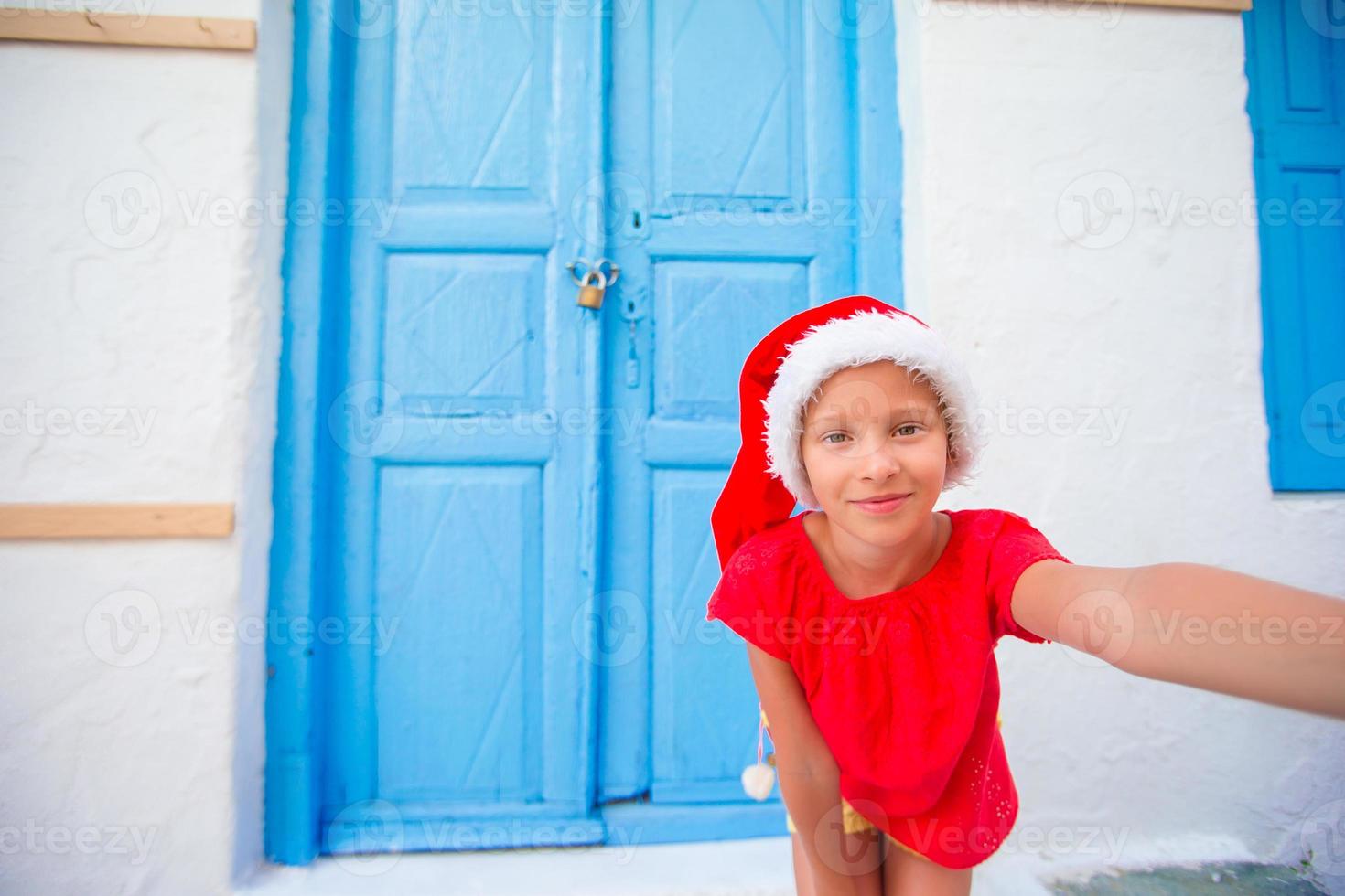 adorável garotinha com chapéu de Papai Noel tomando selfie na rua estreita na vila grega foto