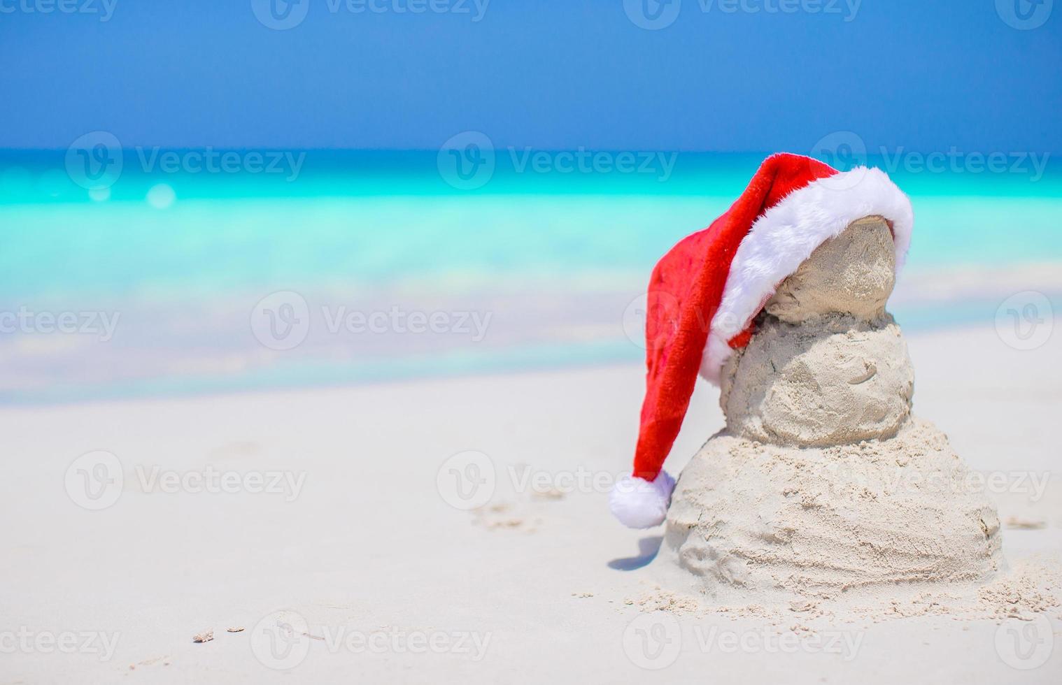 pequeno boneco de neve de areia com chapéu de papai noel vermelho na praia branca do caribe foto