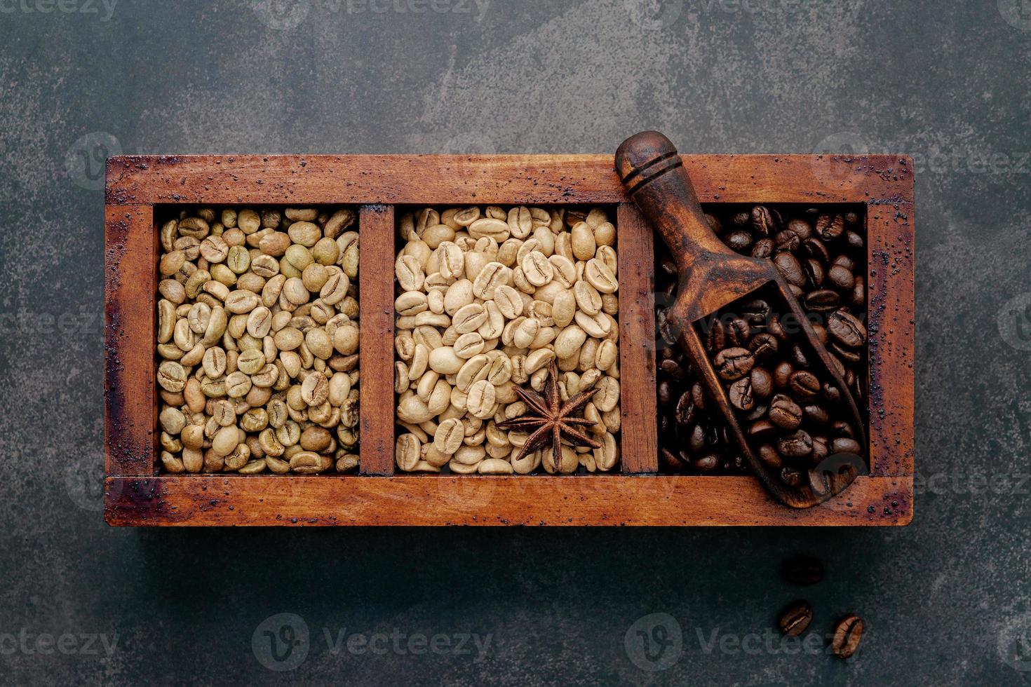 grãos de café torrados e escuros verdes e marrons em caixa de madeira com configuração de colheres em fundo escuro de concreto. foto