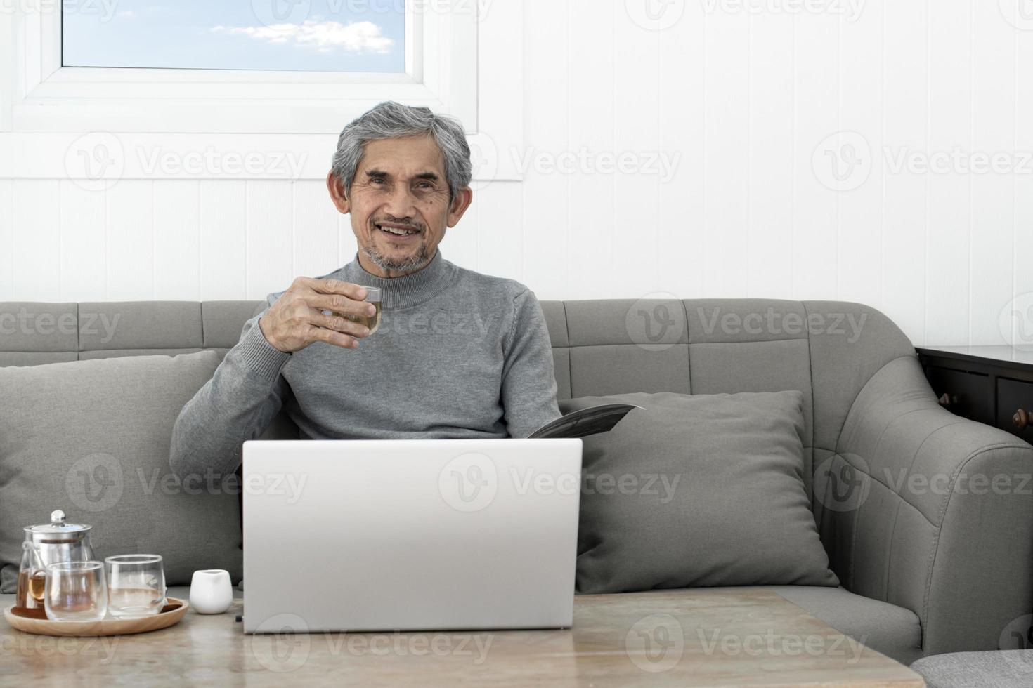 homem idoso de cabelos grisalhos asiáticos com camisa de manga comprida senta-se no sofá e bebe chá quente na sala de estar de sua casa, conceito de vida feliz dos idosos e seguro de vida em todo o mundo. foto