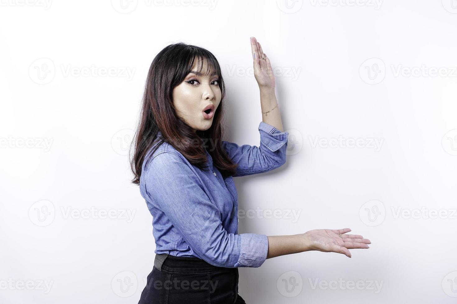 mulher asiática chocada vestindo camisa azul apontando para o espaço da cópia ao lado dela, isolado pelo fundo branco foto