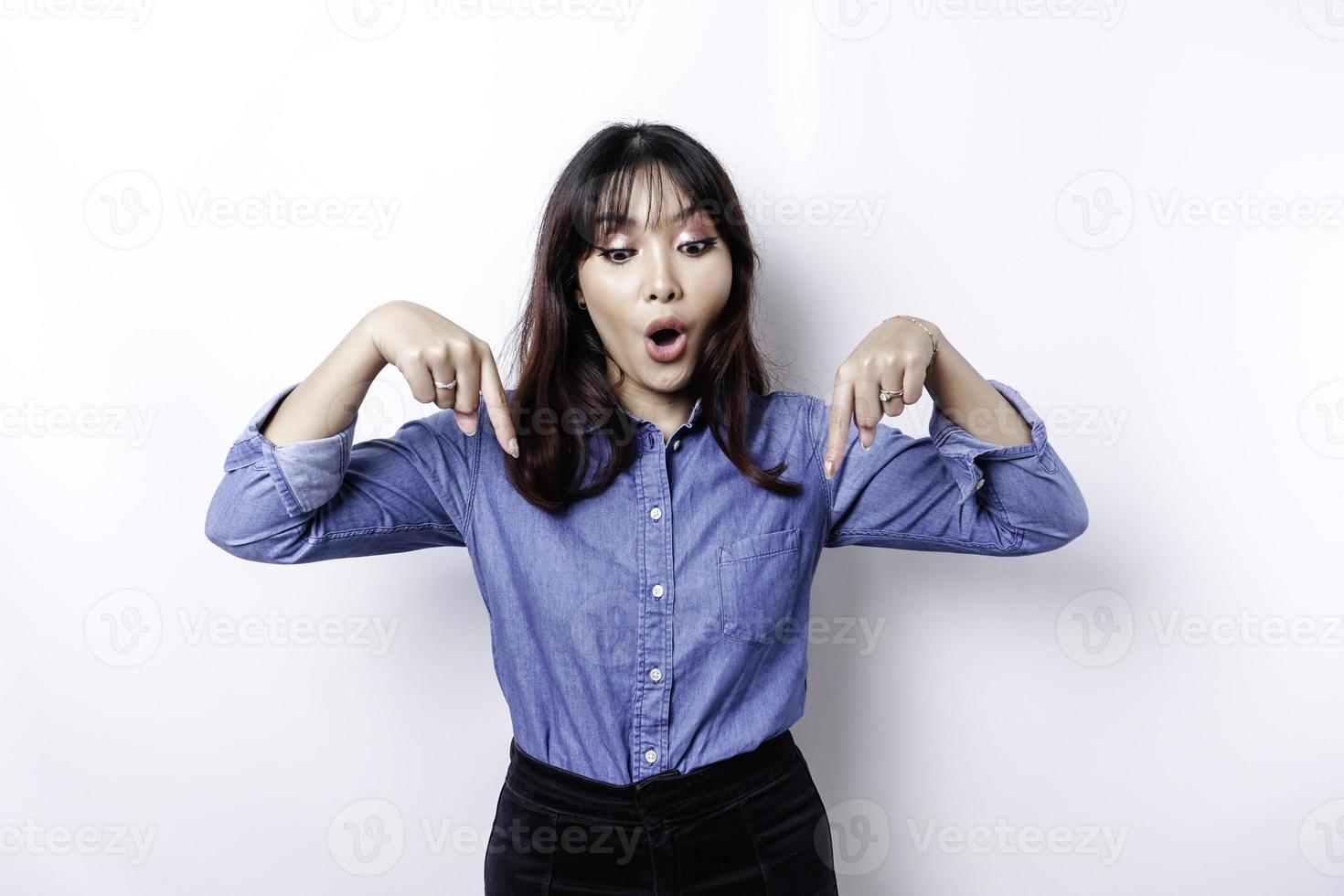 mulher asiática chocada vestindo camisa azul apontando para o espaço da cópia abaixo dela, isolado pelo fundo branco foto