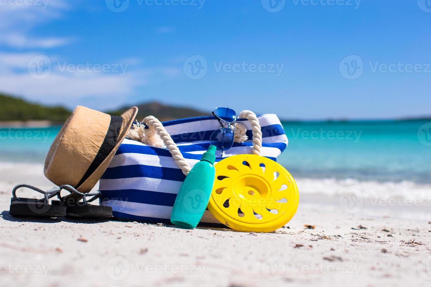 bolsa listrada, chapéu de palha, protetor solar e frisbee na praia tropical de areia branca foto