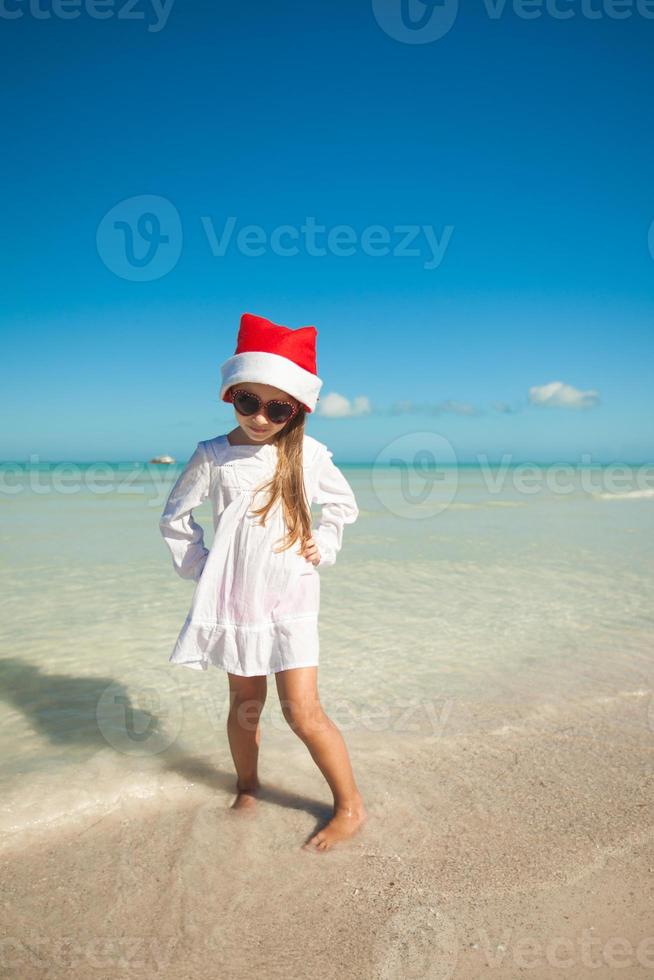 menina bonitinha de chapéu vermelho de papai noel e óculos de sol na praia foto