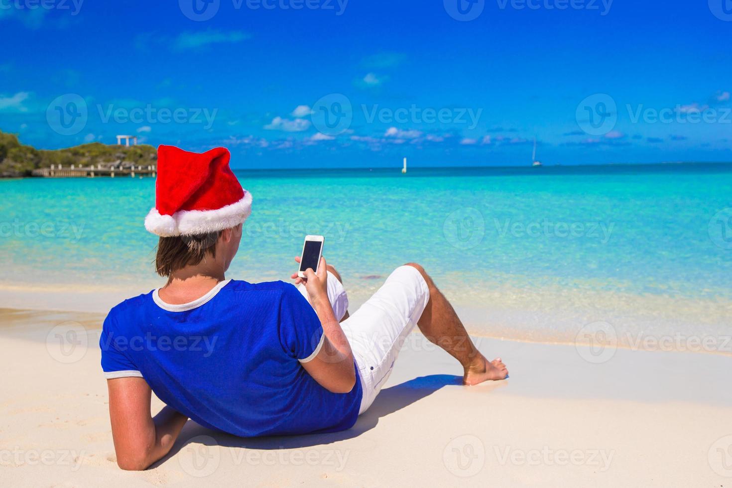 jovem de chapéu de Papai Noel falando no telefone na praia tropical foto