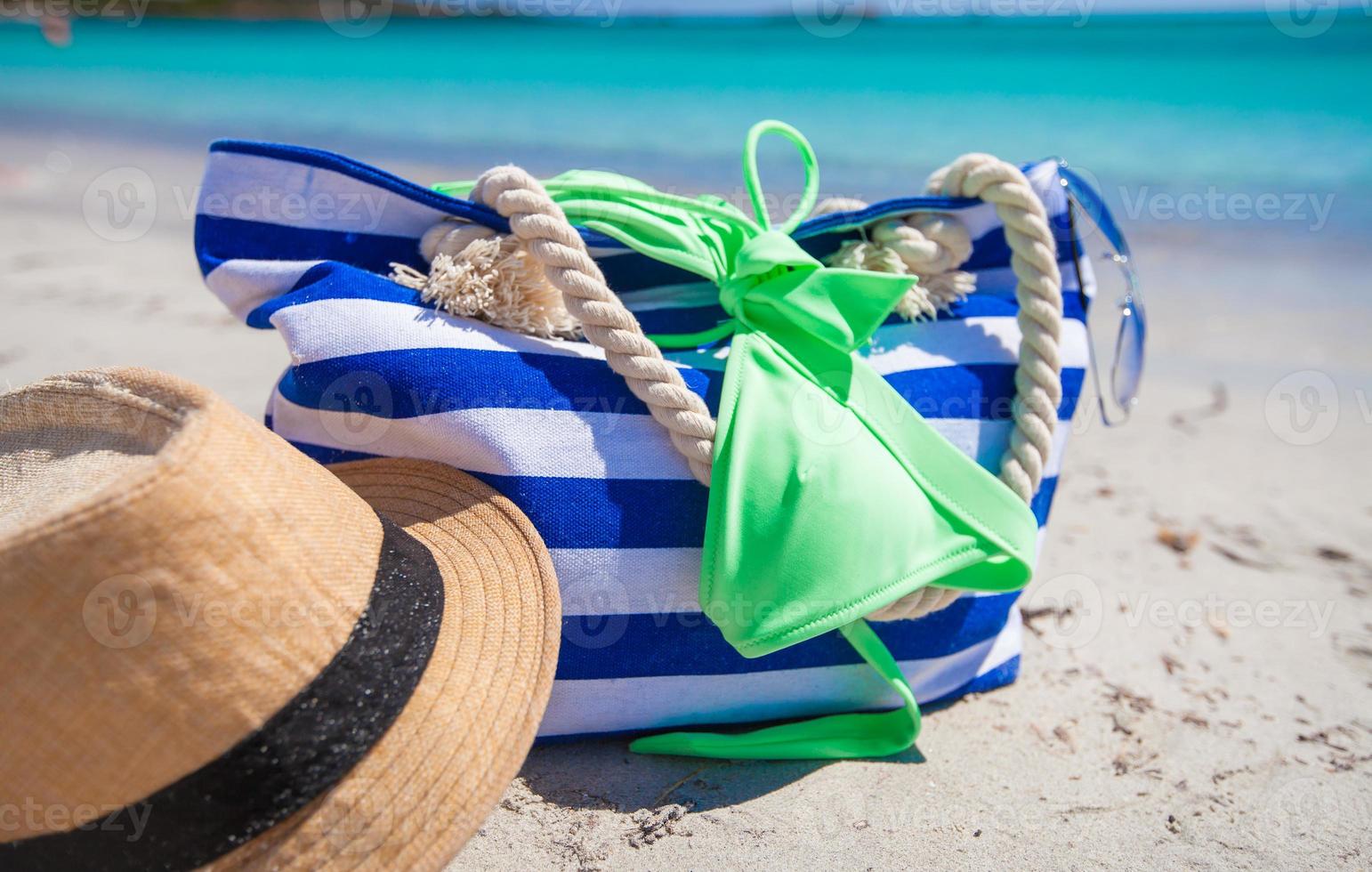 bolsa listrada, chapéu de palha, protetor solar e frisbee na praia tropical de areia branca foto