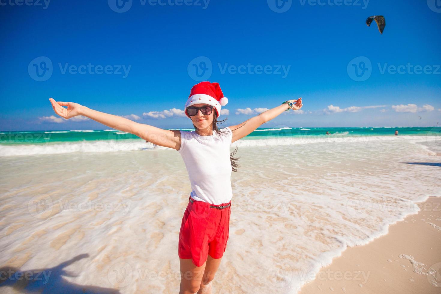 jovem de chapéu de papai noel andando espalhou as mãos na praia de areia branca foto