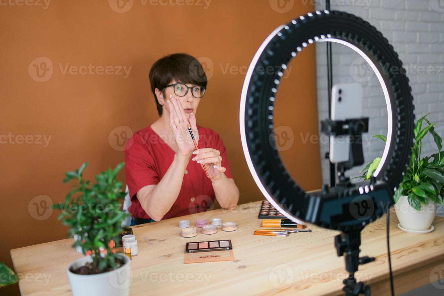 blogueira de beleza feminina de meia-idade ou influenciadora com vídeo de gravação de produto de maquiagem no quarto em casa. usando lâmpada de anel e smartphone foto