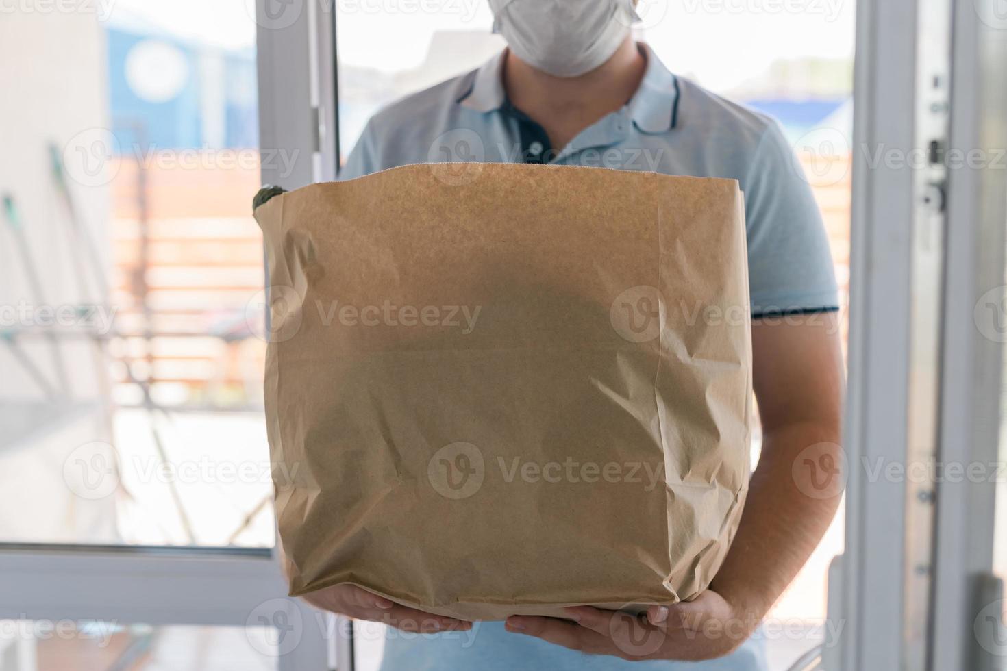 entregar homem usando máscara facial em uniforme azul manuseando saco de comida entregue ao cliente na frente da casa. carteiro e serviço de entrega expressa de supermercado foto