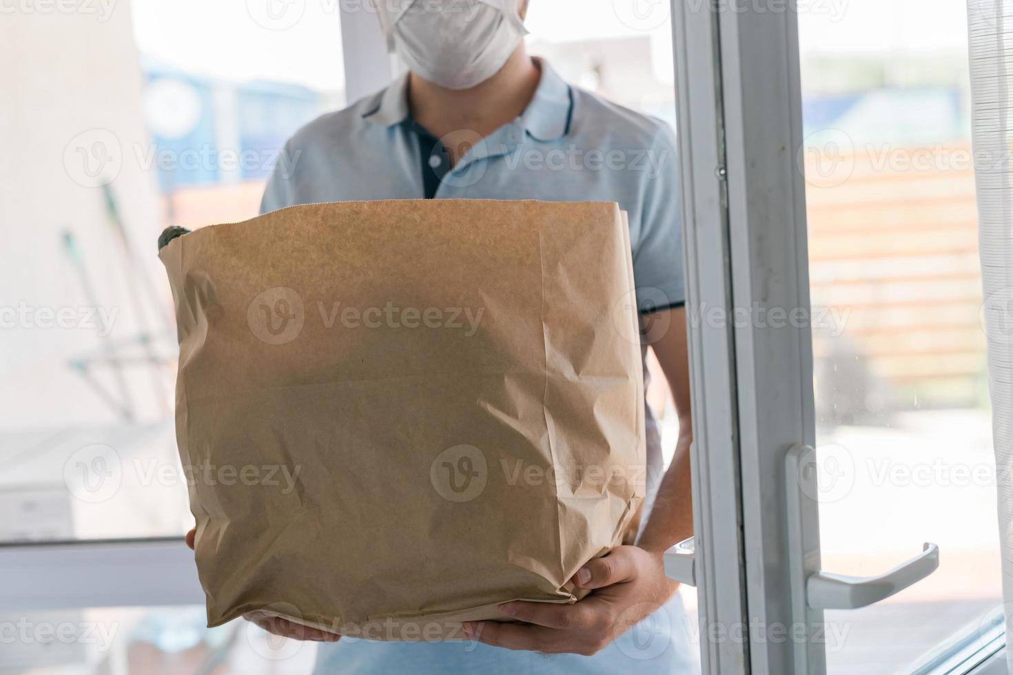 entregar homem usando máscara facial em uniforme azul manuseando saco de comida entregue ao cliente na frente da casa. carteiro e serviço de entrega expressa de supermercado foto