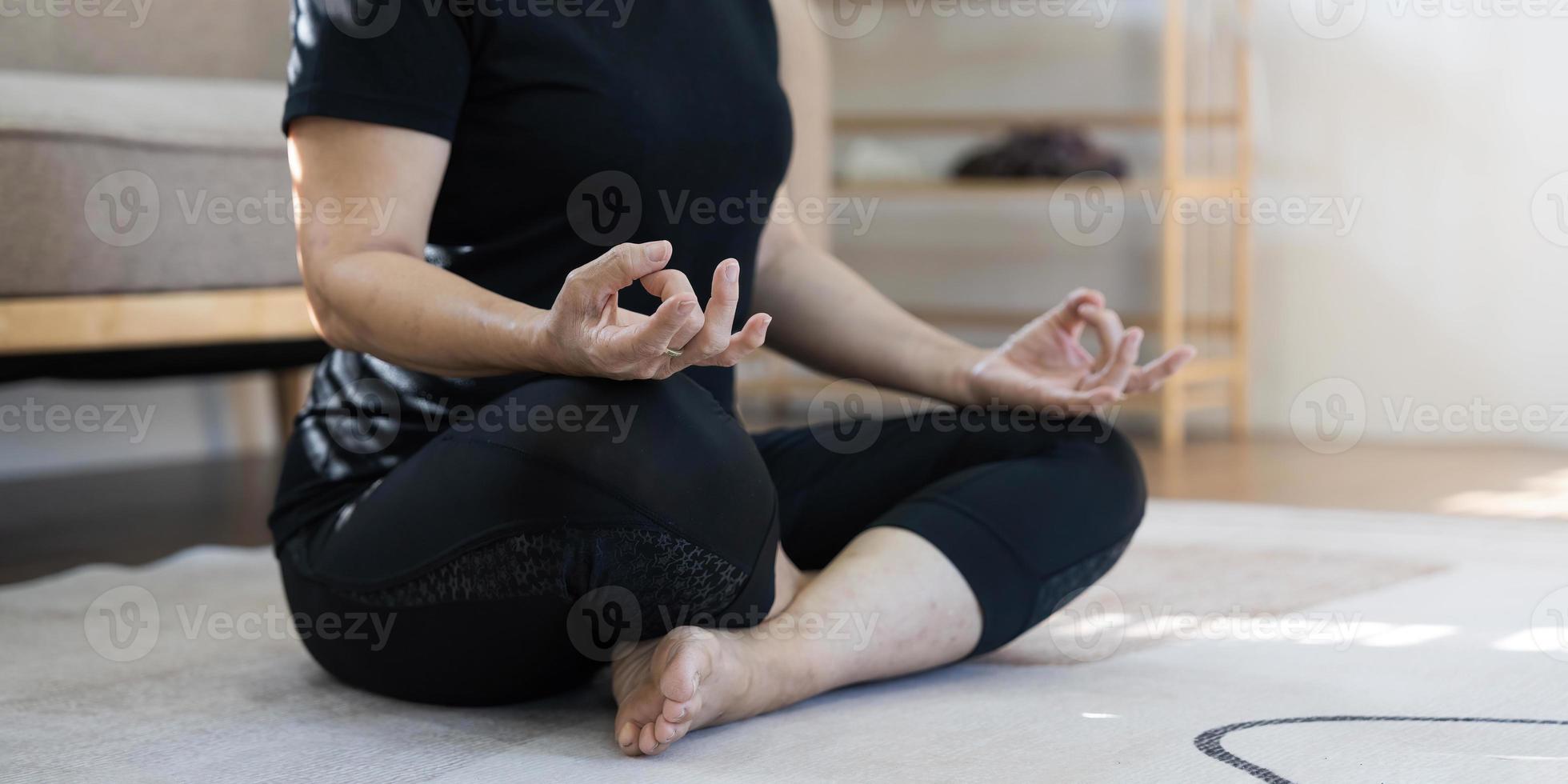 calma e relaxada mulher asiática aposentada dos anos 60 em roupas de ginástica está meditando em sua sala de estar, olhos fechados, praticando pose de ioga de lótus em casa. foto