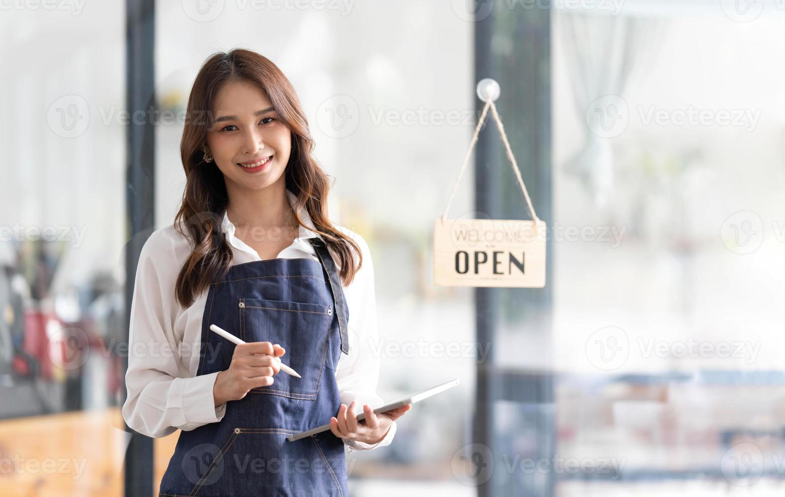 bela jovem asiática barista no avental segurando o tablet e em frente à porta do café com placa aberta. conceito de inicialização do proprietário do negócio. foto