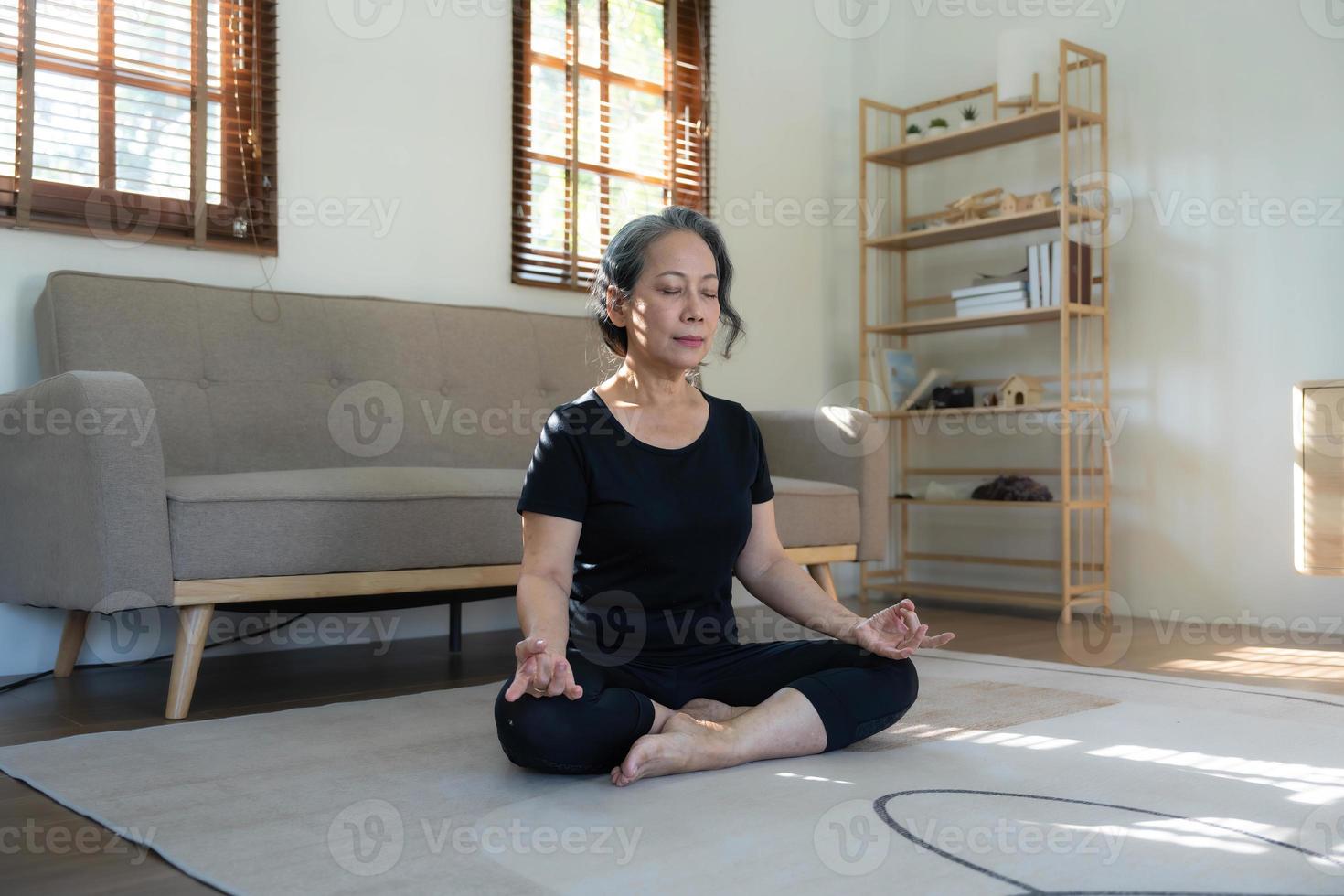calma e relaxada mulher asiática aposentada dos anos 60 em roupas de ginástica está meditando em sua sala de estar, olhos fechados, praticando pose de ioga de lótus em casa. foto