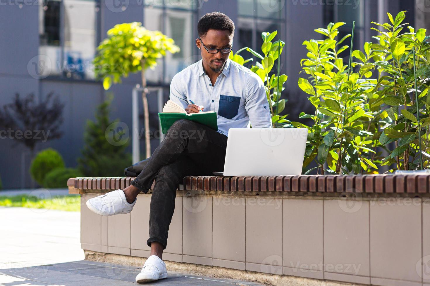 jovem estudante afro-americano sentado em um parque da cidade em um banco com laptop e notebook foto