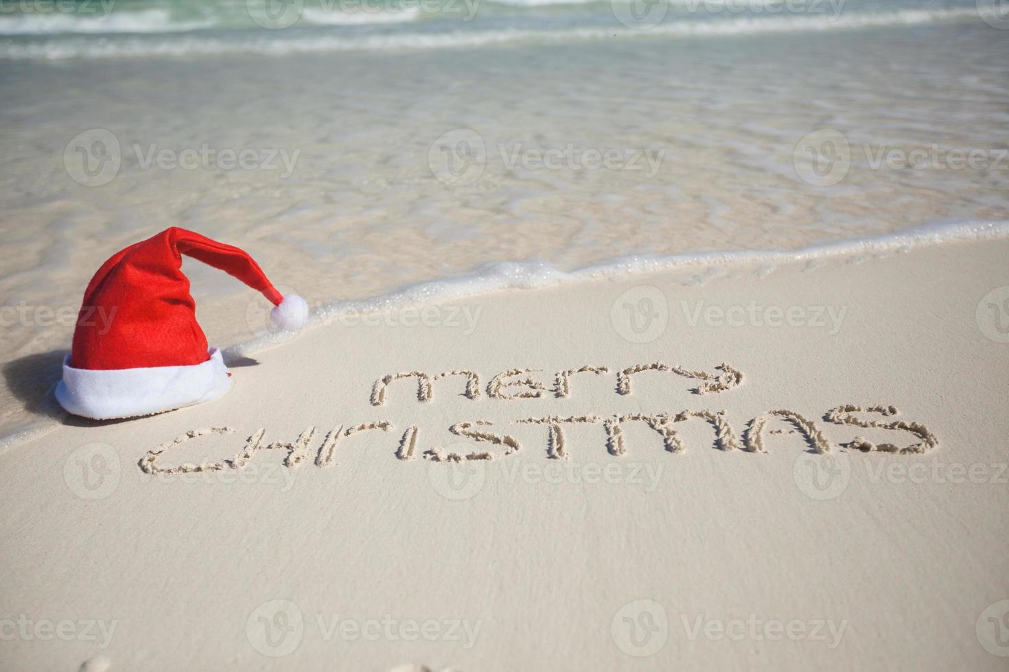 feliz natal escrito na areia branca da praia tropical com chapéu de natal foto