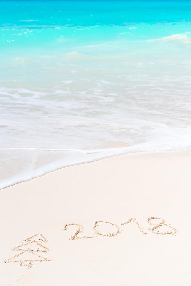 2018 escrito à mão na praia de areia com onda do mar suave no fundo foto