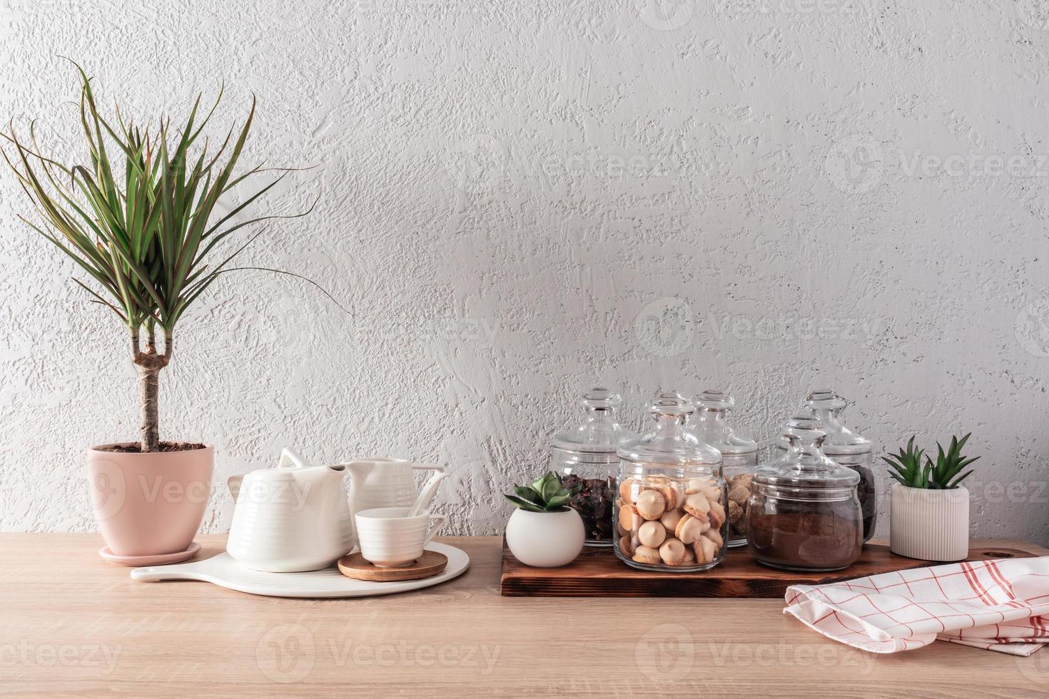 interior da cozinha, plano de fundo. frascos de vidro com chá seco, biscoitos, grãos de café, bule de cerâmica branca e xícaras. plantas. vista frontal. foto