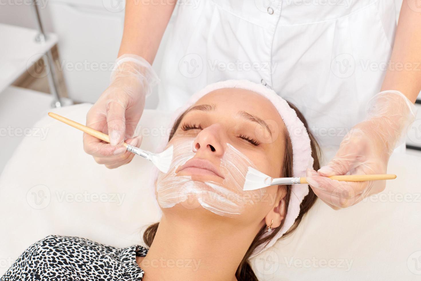 esteticista aplicando máscara de creme cosmético no rosto de mulher para rejuvenescimento, procedimento em salão de beleza foto