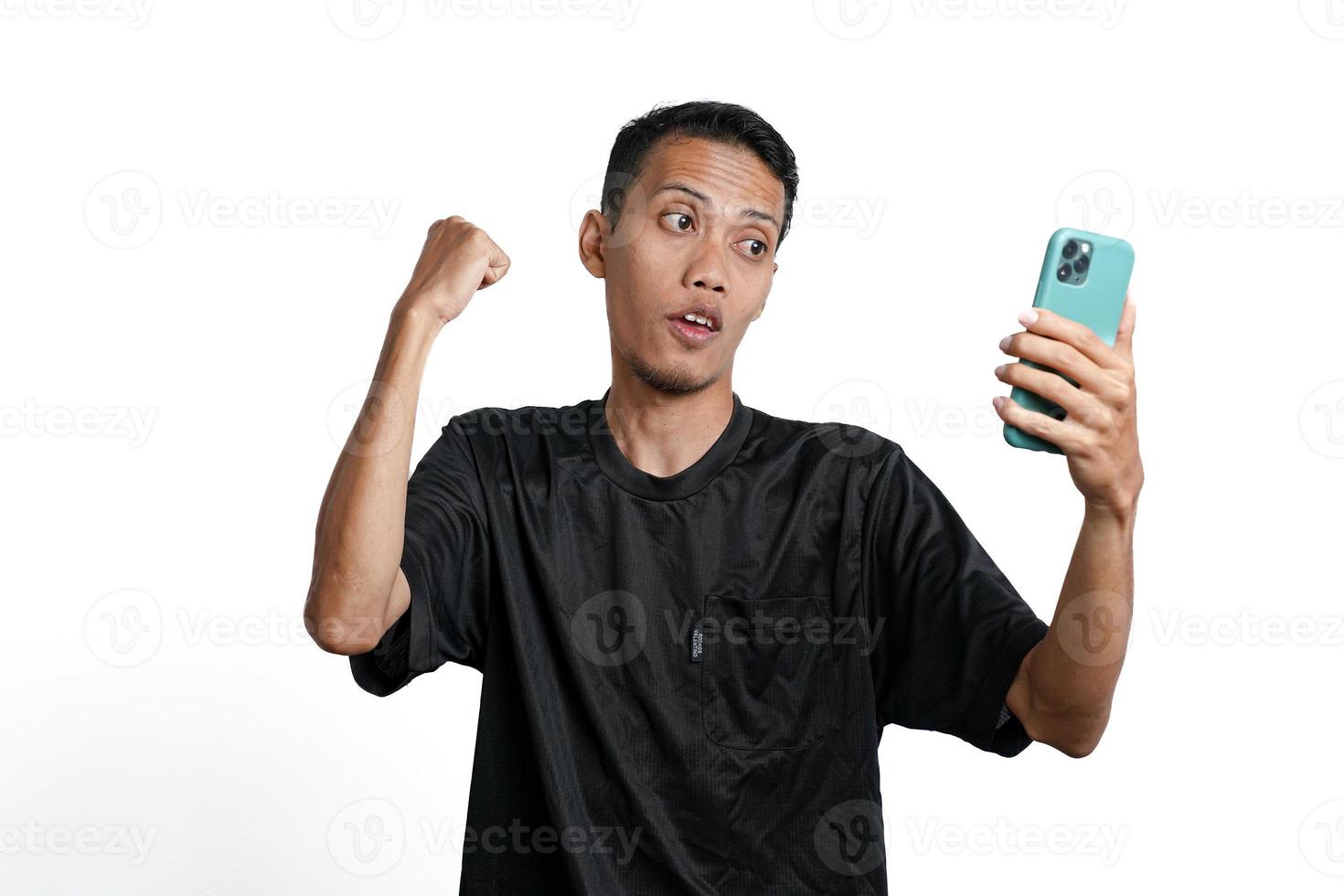 homem asiático vestindo camiseta preta de treinamento, com um gesto feliz e animado enquanto olha para a tela do smartphone. isolado por fundo branco foto
