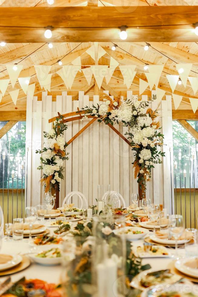 mesa festiva com refeições para casamento, arco de madeira, stands decorados com composição de flores brancas e vegetação no salão de banquetes rústico. foto