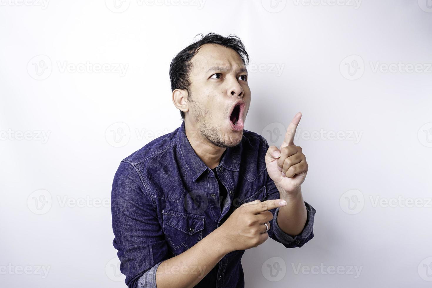 homem asiático chocado vestindo camisa azul apontando para o espaço da cópia ao lado dele, isolado pelo fundo branco foto