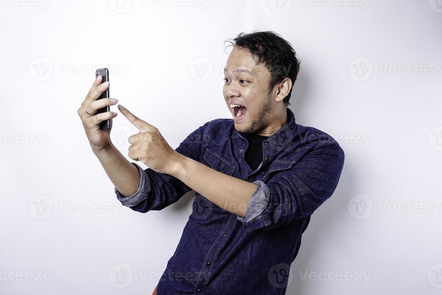 homem asiático chocado vestindo camisa azul e segurando seu telefone, isolado por fundo branco foto