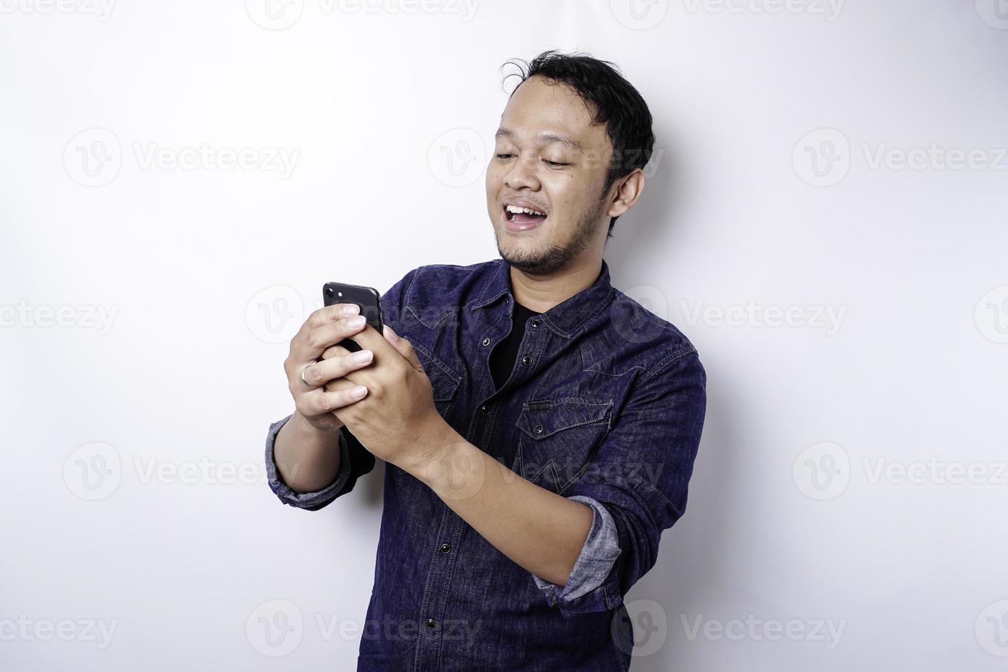 homem asiático animado vestindo camisa azul sorrindo enquanto segura seu telefone, isolado por fundo branco foto