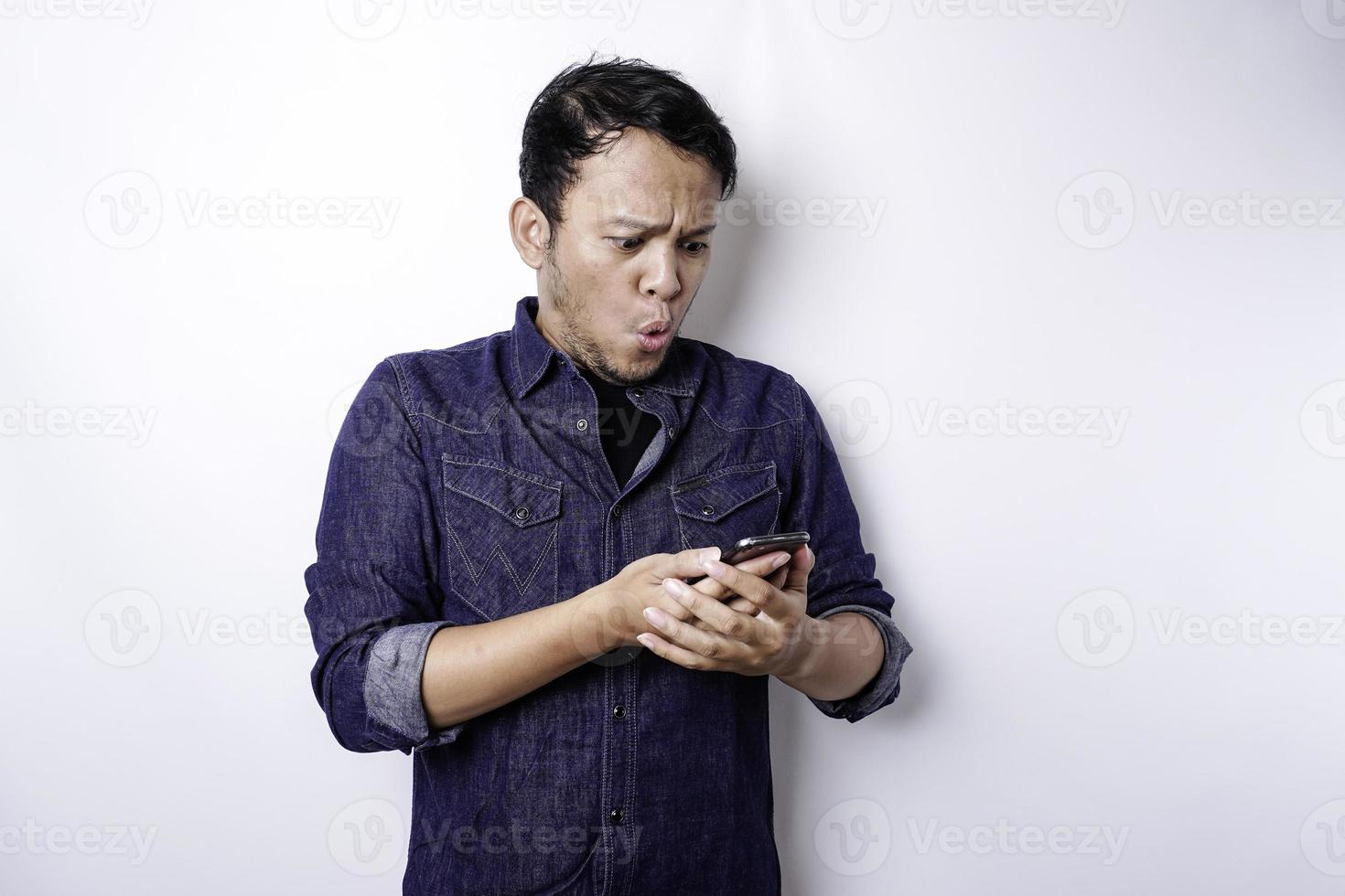 homem asiático chocado vestindo camisa azul e segurando seu telefone, isolado por fundo branco foto