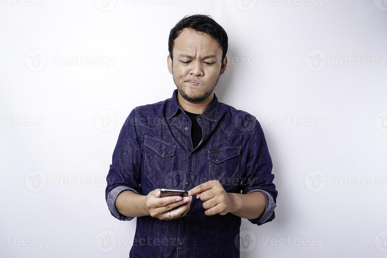 um jovem asiático pensativo está vestindo camisa azul segurando seu telefone e parece confuso, isolado pelo fundo branco foto